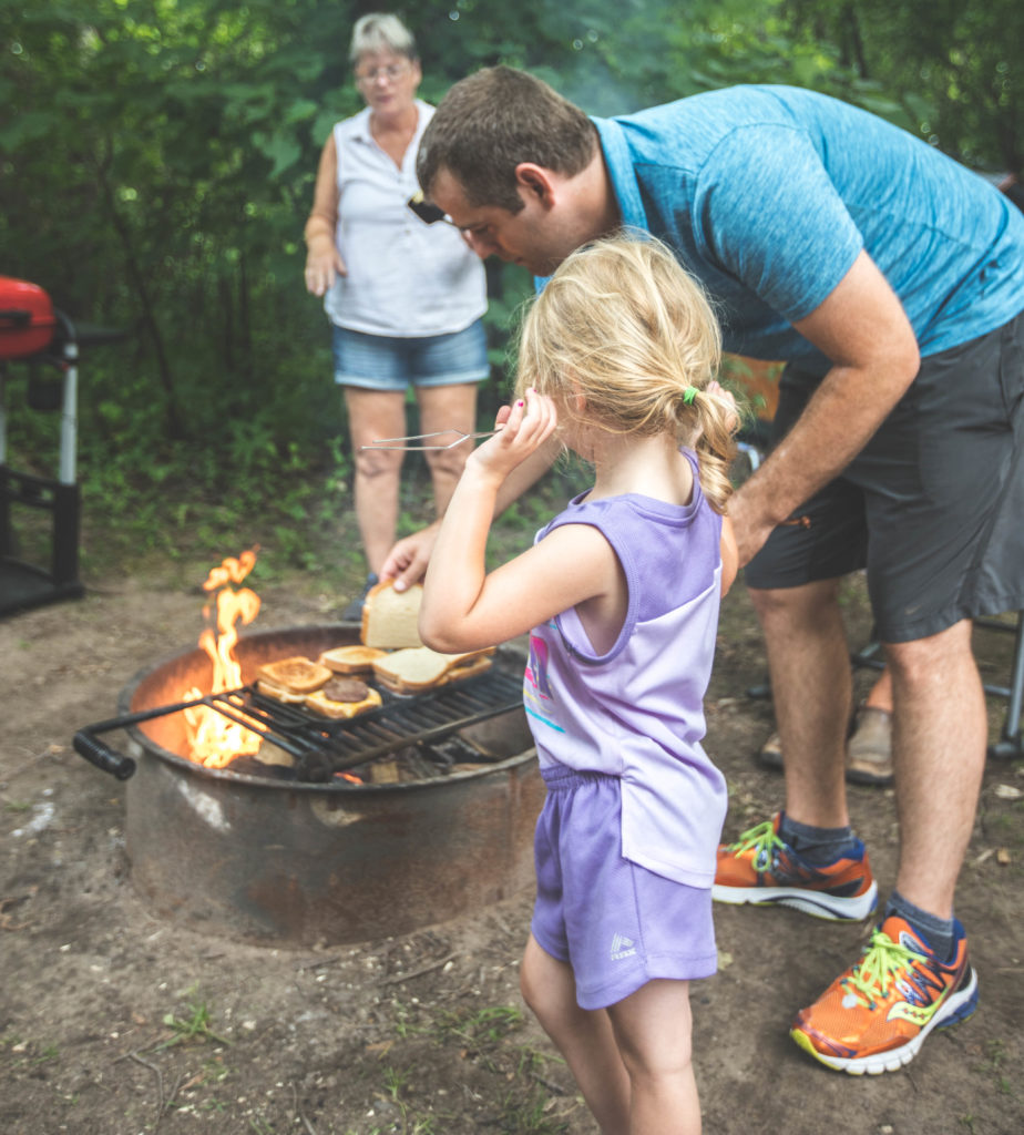 4 year old helping dad cook burgers over campfire with tent camping